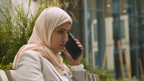 Muslim-Businesswoman-Answering-Call-On-Mobile-Phone-Sitting-Outside-Office-In-City-Gardens